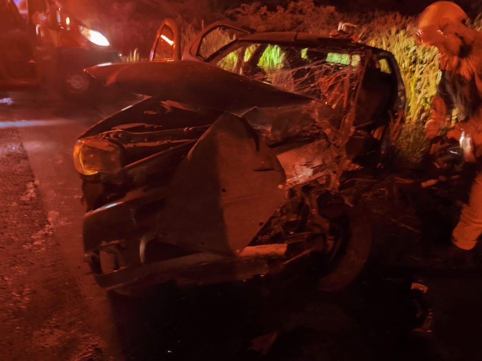 Foto mostra carro destruído após colisão contra caminhão-tanque. O veículo Siena tem cor cinza e a foto foi tirada à noite