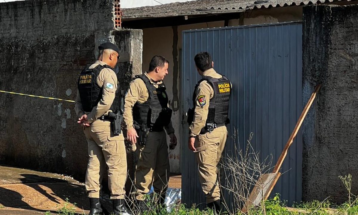 Foto tirada durante o dia mostra três policiais militares fardados parados em frente a uma casa, ao lado de um portão de ferro grande. No chão, dentro do terreno, há um corpo coberto por uma manta de alumínio