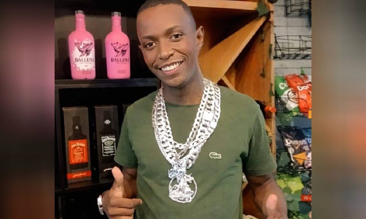Foto mostra homem com colar de prata no pescoço e garrafas de bebidas ao fundo