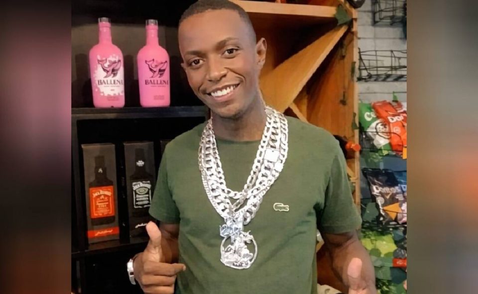 Foto mostra homem com colar de prata no pescoço e garrafas de bebidas ao fundo