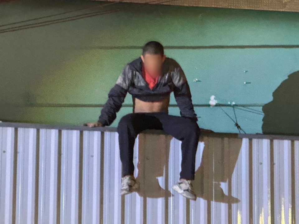 Homem aparece sentado em cima de marquise. Ele tem cabelos quase raspados, usa calça preta e jaqueta