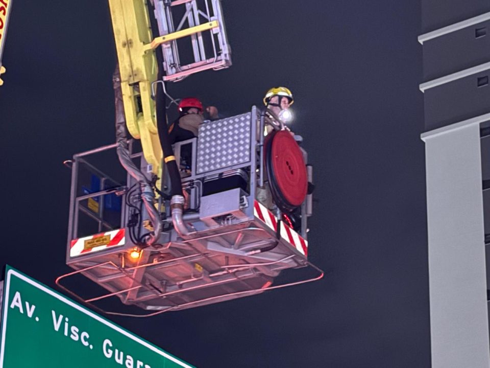 Bombeiro e policial militar aparecem em cima de plataforma mecância do Corpo de Bombeiros. Foto foi tirada à noite.