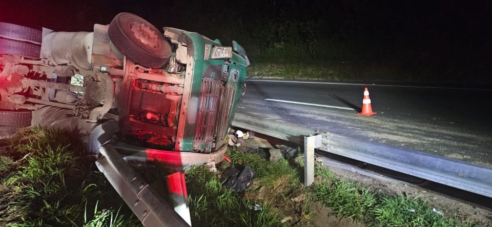 Foto tirada à noite mostra cabine de caminhão verde de ponta cabela após tombar às margens de rodovia