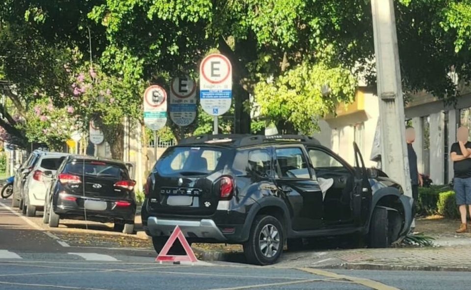 Foto mostra um veículo Renault Duster preto parado após bater contra um poste em um cruzamento do bairo Batel, em Curitiba. Atrás do carro há um triângulo de sinalização.