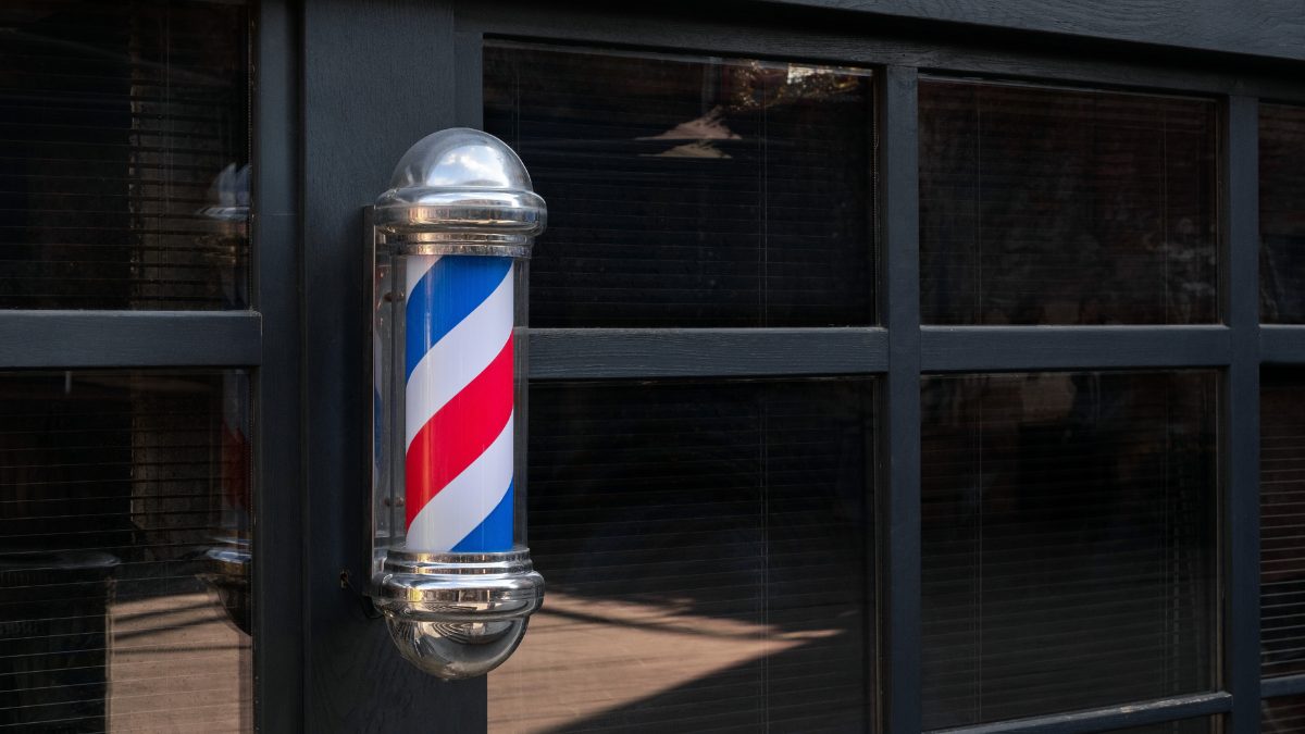 Foto ilustrativa mostra poste de barbearia, um cilindro giratório com espiral nas cores vermelha, branca e azul, instalado na parede de estabelecimento. Uma janela escura aparece na foto