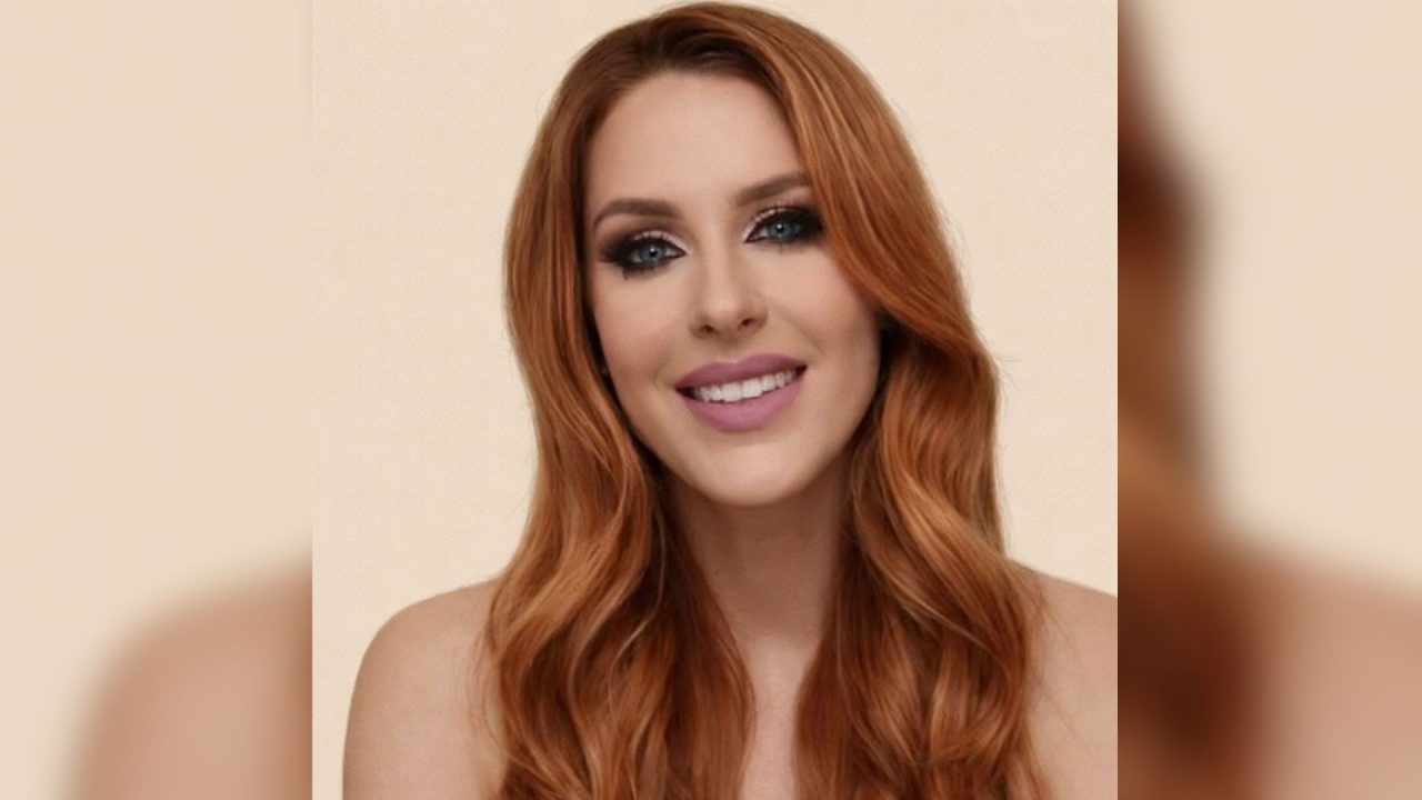 Foto em estilo 3x4 mostra Adriana Ligoski de Campos Barreto olhando para a câmera e sorrindo. Ela usa cabelos ruivos, batom claro e maquiagem escura nos olhos. Ela tem a pele branca.