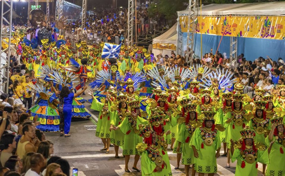 Foto mostra desfile de escola de samba com pessoas fantasiadas e o público assistindo