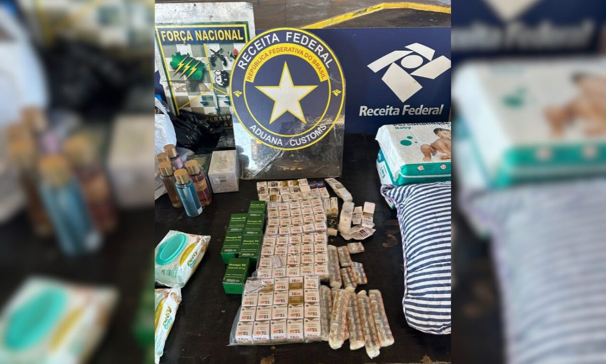 Mesa com medicamentos apreendidos expostos em frente a placas da Receita Federal e da Força Nacional. Entre os itens estão diversas caixas pequenas organizadas em fileiras, ampolas embaladas em plástico, frascos de cosméticos, pacotes de lenços umedecidos, fraldas e uma bolsa listrada.