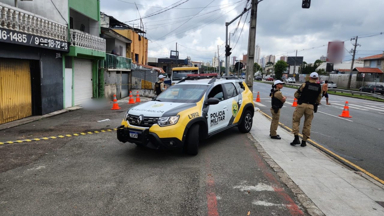 Imagem mostra viatura da Polícia Militar (PM) estacionada em frente a comércios no endereço da ocorrência. O corpo da vítima aparece coberto por um lençol branco, mas borrado via edição.