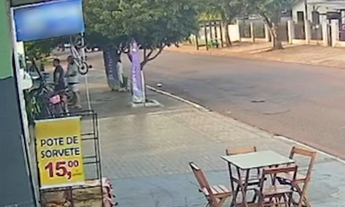 Foto registrada por câmera de segurança mostra dois homens à esquerda da tela, na frente da porta de um comércio. Próximo a ele, em primeiro plano, há uma sorveteria com mesa na calçada e uma placa amarela. Está de dia e os dois homens estão próximos a uma árvore de grande porte