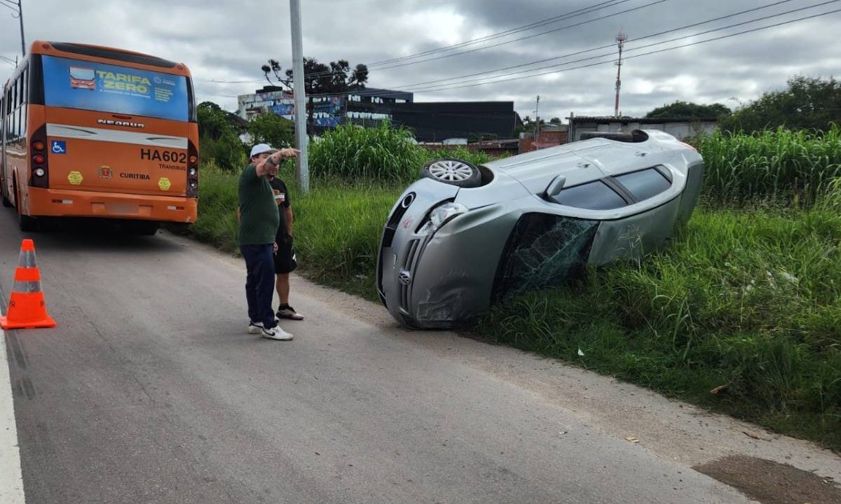 Foto mostra carro tombado em matagal enquanto dois homens acompanhando desdobramentos do caso. À frente, um ônibus laranja aparece estacionado às margens da rua