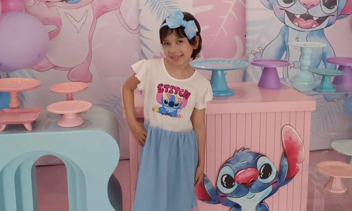 A menina Júlia Soares Teixeira da Silva posa para foto durante a própria festa de aniversário de sete anos. A comemoração foi feita inspirada no filme Stitch, que aparece desenhado em uma mesa. Júlia usa camisa com o rosto de Stitch estampado, saia azul e tiara azul.