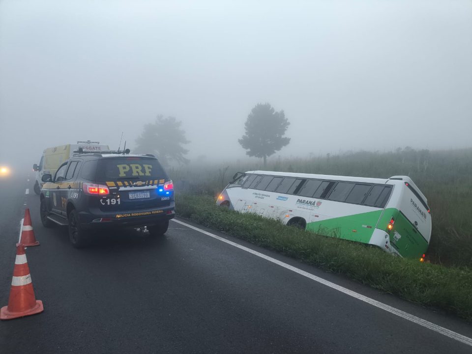 Foto tirada por volta das 5h da manhã mostra ônibus de Secretaria da Saúde, branco com verde, tombado às margens de rodovia, sobre grama. Uma viatura da PRF está estacionada a poucos metros de distância