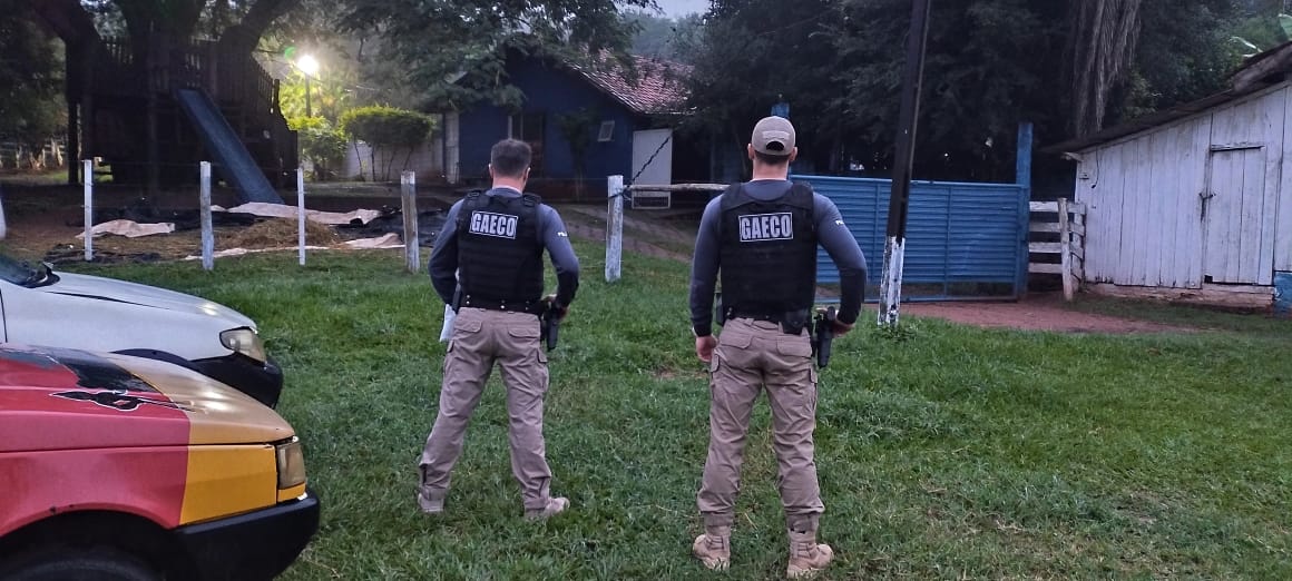 Foto mostra dois agentes do Gaeco de costas, olhando para casa de madeira ao fundo. Eles estão de pé sobre grama e fardados, com colete à prova de balas