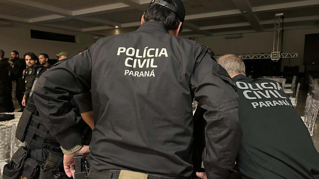 Foto mostra policiais civis fardados de costas para câmera e levemente inclinados para baixo, como se estivessem analisando algo sobre uma mesa