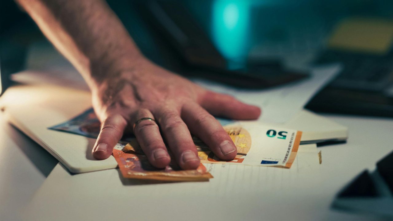 Foto ilustrativa mostra mão de homem sobre cédulas de dinheiro, em cima de uma mesa