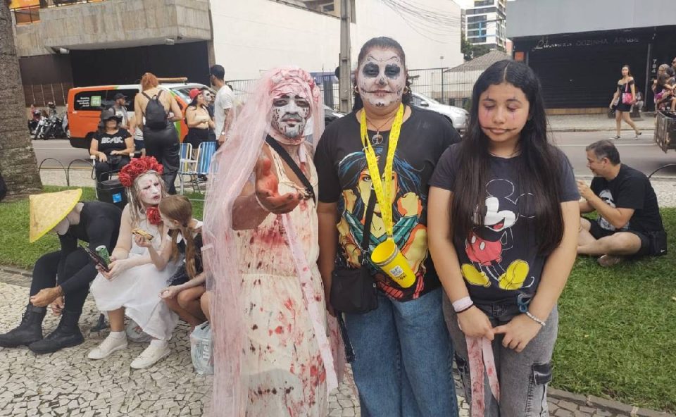 Foto mostra grupo com o rosto pintado de caveira mexicana e homem com vestido de noiva zombie