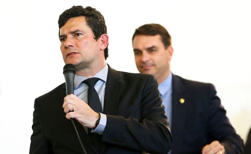O senador Sérgio Moro fala ao microfone em primeiro plano, vestindo terno e gravata escuros. Ao fundo, levemente desfocado, o senador Flávio Bolsonaro observa sorrindo