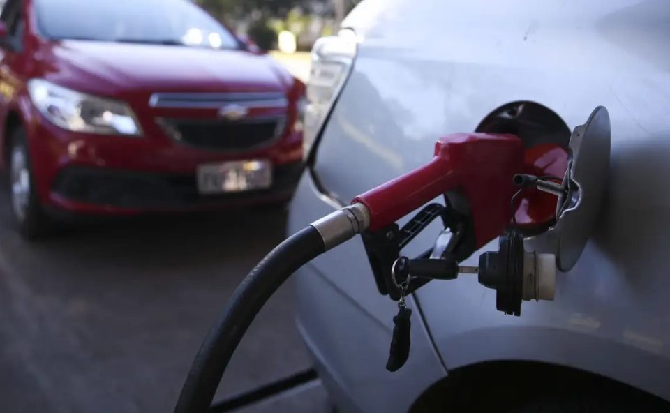 Bomba de combustível abastecendo tanque de um carro prata. ao fundo, há um carro vermelho