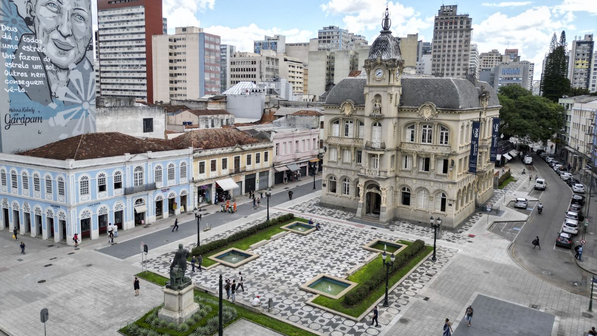Vista aérea da Praça Generoso Marques em Curitiba, destacando o prédio histórico do Paço da Liberdade com sua arquitetura neoclássica e o mosaico de petit-pavé no chão.