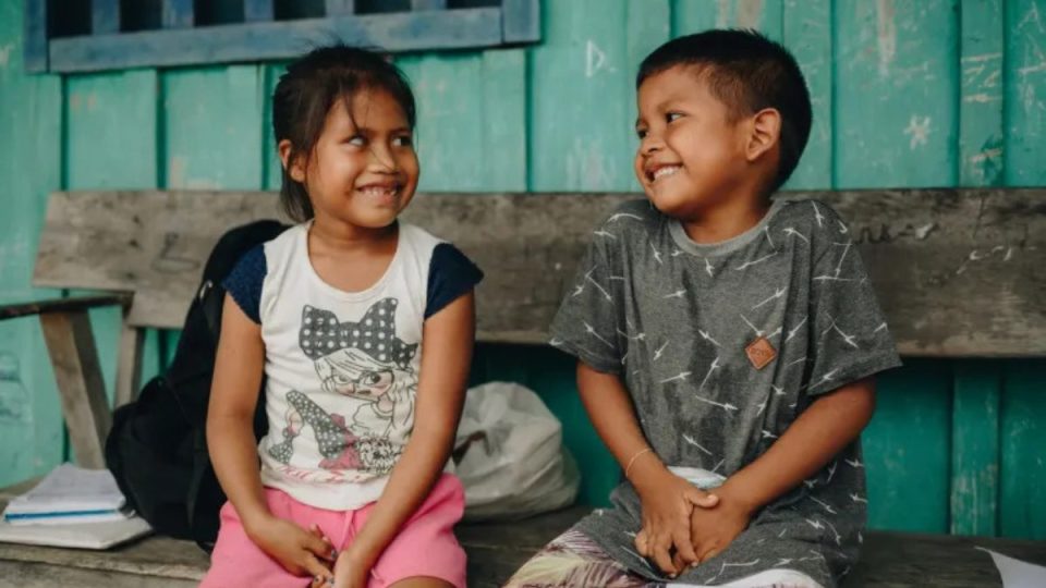 Duas crianças indígenas sorriem sentadas em um banco de madeira em frente a uma escola rural. Ao lado, uma mochila e cadernos, simbolizando o direito à educação e infância protegida no Brasil