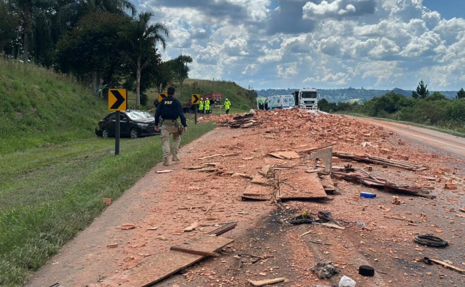 Imagem mostra a carga de tijolos que caiu de um caminhão e provocou capotamento de um carro na BR-277. Foto mostra a carga caída e espalhada na rodovia. Um policial está ao lado da carga.