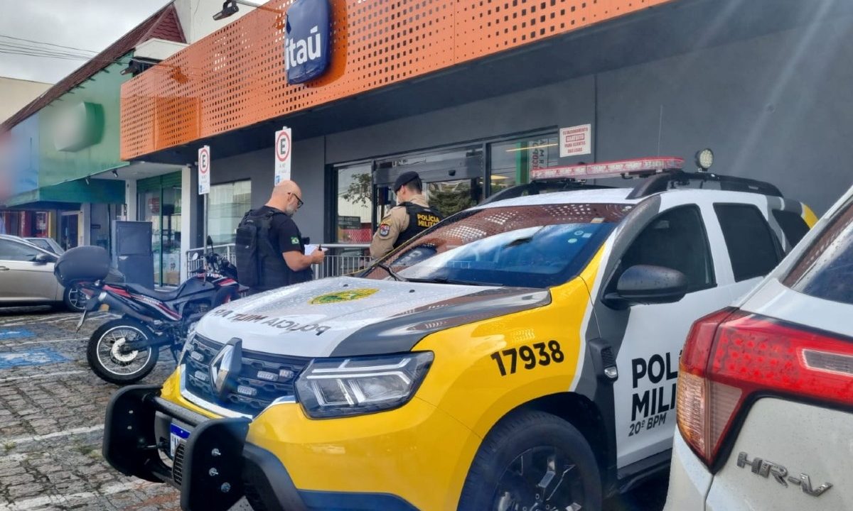 Foto mostra viatura da Polícia Militar parada em frente à agência bancária do Itaú que foi arrombada no bairro Boqueirão, em Curitiba; policiais civil e militar conversam ao lado da viatura