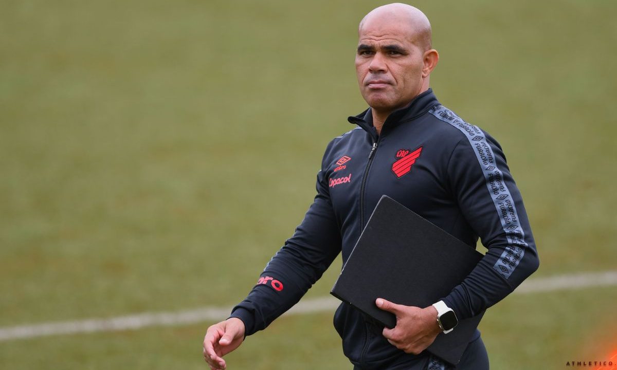 Alessandro, ex-lateral e campeão brasileiro pelo Athletico, aparece caminhando à beira de um campo de treinamento no CT do Caju. Ele veste agasalho preto do clube com o escudo rubro-negro no peito e segura uma prancheta enquanto observa a atividade, durante período em que trabalhou nas categorias de base.