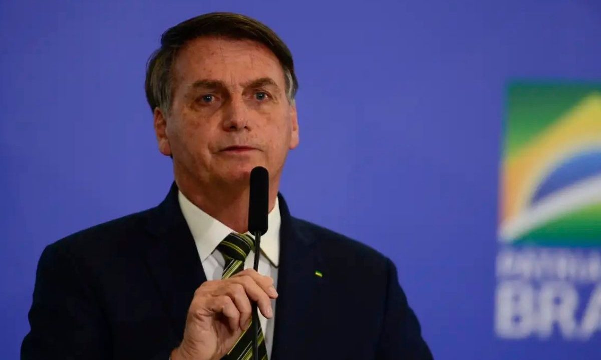 A imagem mostra Jair Bolsonaro de terno preto, falando ao microfone, e, ao fundo, uma imagem azul com a logo do governo federal da época em que ele era presidente.
