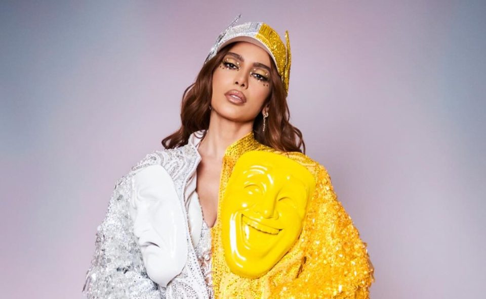 Imagem mostra a cantora Anitta, que apresentará o SNL, em ensaio de fotos. Ela usa um boné branco e amarelo, com casaco da mesma cor e com máscaras teatrais.