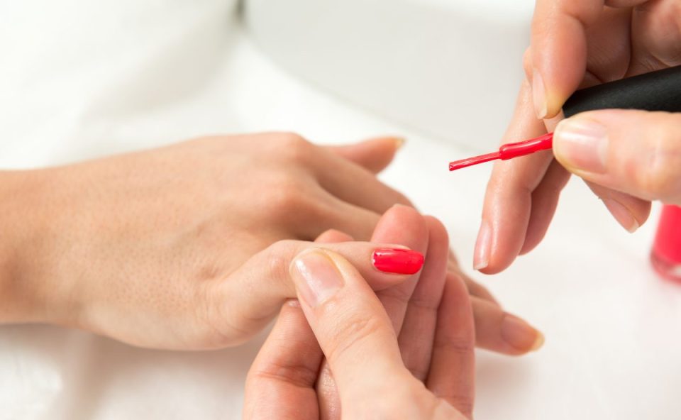 Mãos de uma manicure aplicando esmalte nas unhas de outra pessoa durante atendimento em salão de beleza.
