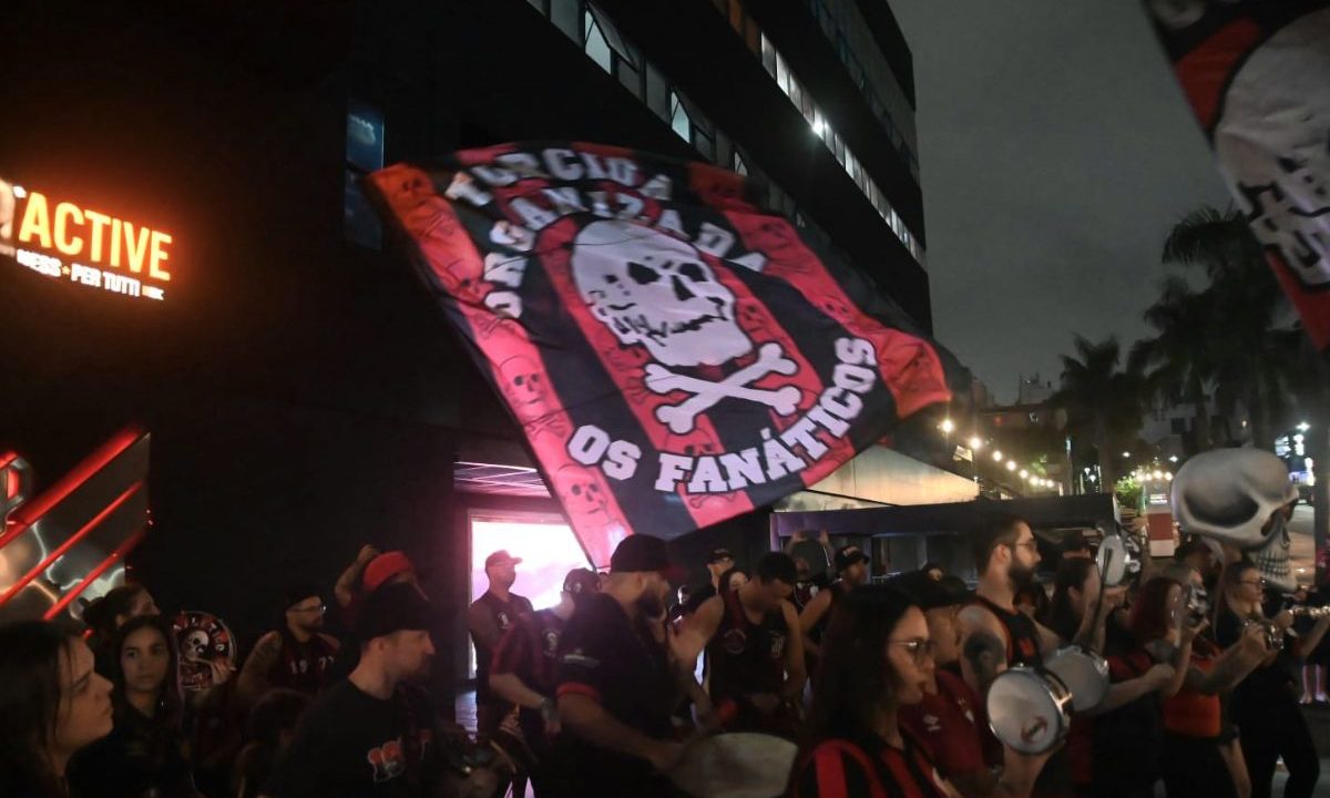 Torcedores do Athletico, com camisetas rubro-negras, tocam instrumentos e seguram uma grande bandeira com símbolo de caveira da organizada Os Fanáticos durante o Aniversário do Athletico, em frente à Arena da Baixada, à noite, com prédios iluminados ao fundo.