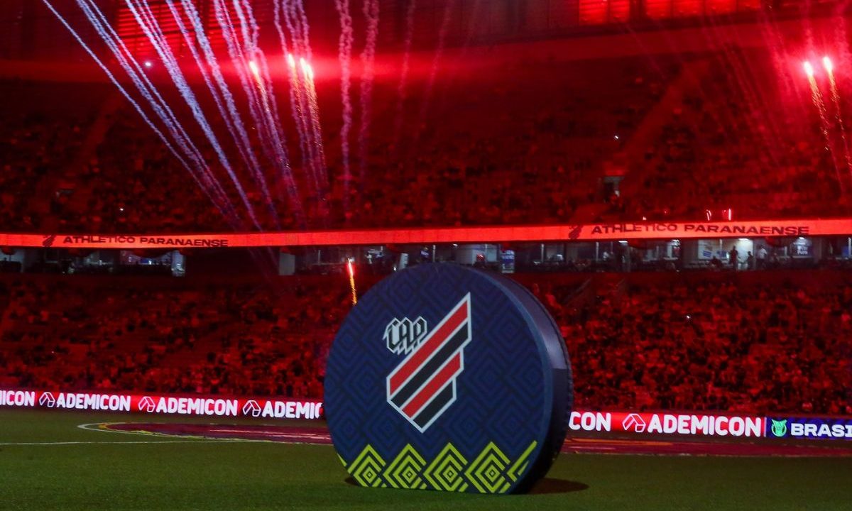 Imagem registrada dentro da Arena da Baixada, iluminada predominantemente em vermelho. No centro do gramado, há um grande painel circular com o escudo do Athletico em destaque. Ao fundo, as arquibancadas estão cheias de torcedores, também banhadas pela iluminação vermelha do estádio. No alto, fogos de artifício são disparados, criando rastros luminosos no ar e reforçando o clima de espetáculo antes da partida. Faixas luminosas ao redor do estádio exibem o nome “Athletico Paranaense”.