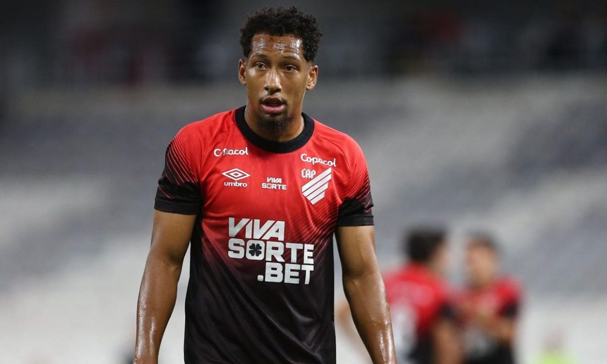 Jogador Habraão, do Athletico, aparece em campo durante partida, vestindo camisa rubro-negra com detalhes em preto, com expressão séria e suor no rosto, enquanto o fundo mostra outros atletas desfocados no estádio