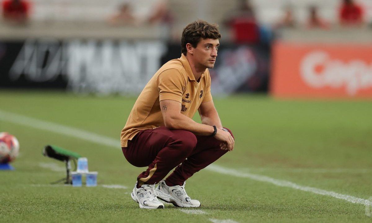 Um homem jovem, de pele clara e cabelos castanhos curtos, está agachado à beira do campo de futebol. Ele veste uma camisa polo dourada com detalhes do Athletico, calça esportiva vermelha e tênis branco. Sua expressão é séria e concentrada, com o olhar fixo para dentro do campo, como se acompanhasse atentamente o jogo ou o treino. Ao fundo, desfocado, aparecem arquibancadas e elementos do estádio, com predominância das cores vermelho e preto. No chão, próximo a ele, há uma garrafa de água e um objeto esportivo, possivelmente parte do material de treino. A cena transmite um momento de observação e análise à beira do gramado.