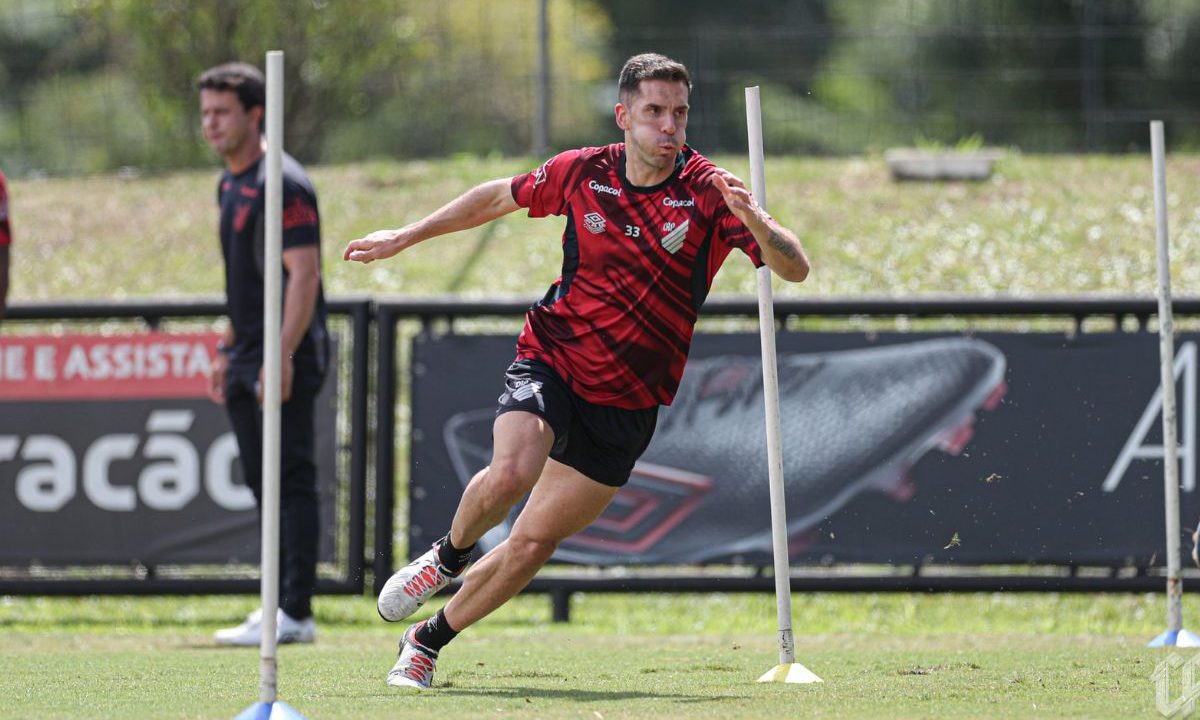 Juan Felipe Aguirre, zagueiro do Athletico realiza treino físico em campo, correndo entre estacas durante atividade de agilidade, vestindo uniforme vermelho e preto do clube, com gramado ao fundo e outro profissional observando.
