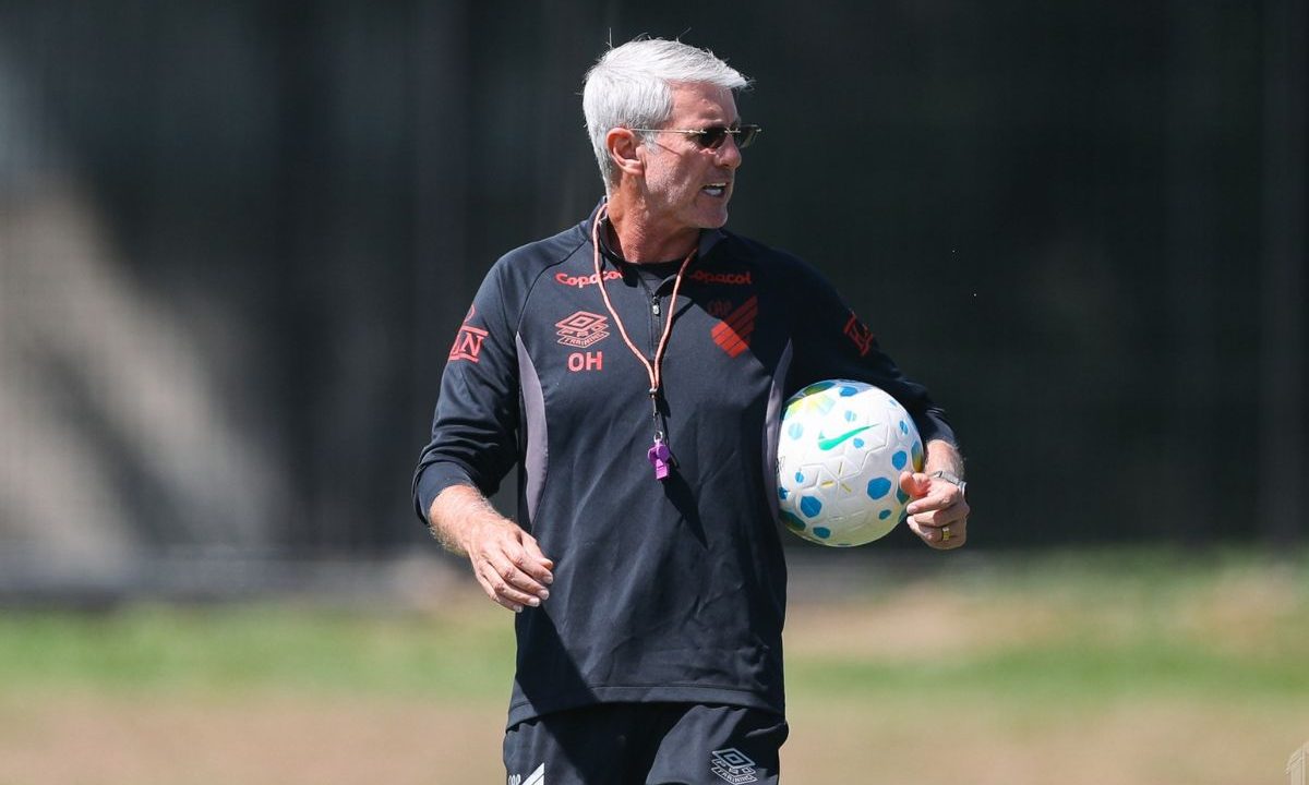Odair Hellmann, técnico do Athletico, aparece durante treino no CT do Caju, vestindo uniforme preto do clube e segurando uma bola, enquanto observa a atividade em campo antes de definir provável escalação do Athletico para enfrentar o Bahia.