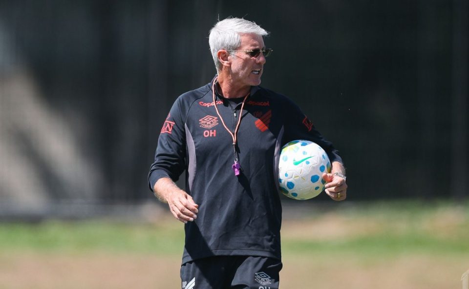Odair Hellmann, técnico do Athletico, aparece durante treino no CT do Caju, vestindo uniforme preto do clube e segurando uma bola, enquanto observa a atividade em campo antes de definir provável escalação do Athletico para enfrentar o Bahia.