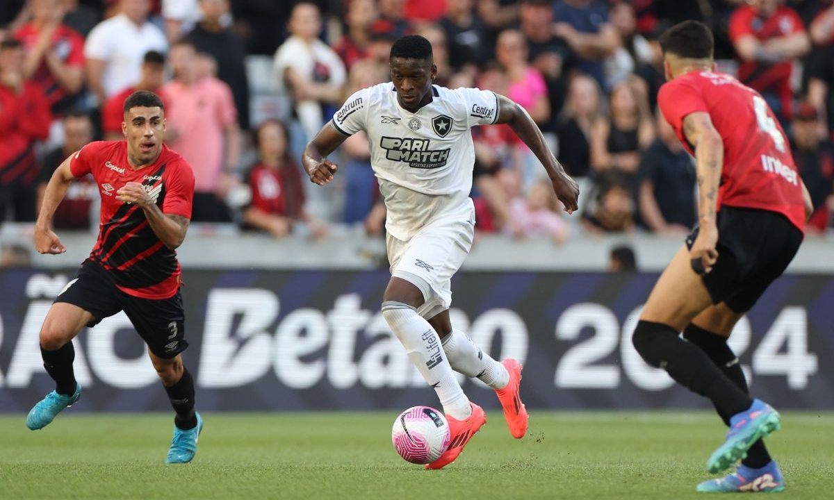Em campo de futebol, um jogador do Botafogo, vestindo uniforme branco e chuteiras rosas, conduz a bola em velocidade pelo gramado. Ele está no centro da imagem, focado na jogada, enquanto dois jogadores do Athletico, com camisas rubro-negras, se aproximam para tentar a marcação — um pela esquerda e outro pela direita. Ao fundo, a arquibancada aparece ocupada por torcedores, muitos com roupas nas cores do Athletico, acompanhando o lance. A cena mostra um momento de transição ofensiva do time carioca sob pressão da defesa adversária.