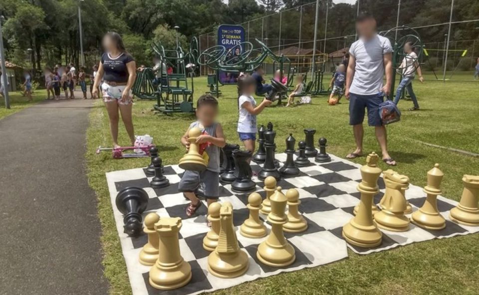Criança e adultos em um parque de Curitiba brincando em um grande tabuleiro de xadrez, uma das atividades gratuitas disponibilizadas pela Prefeitura.