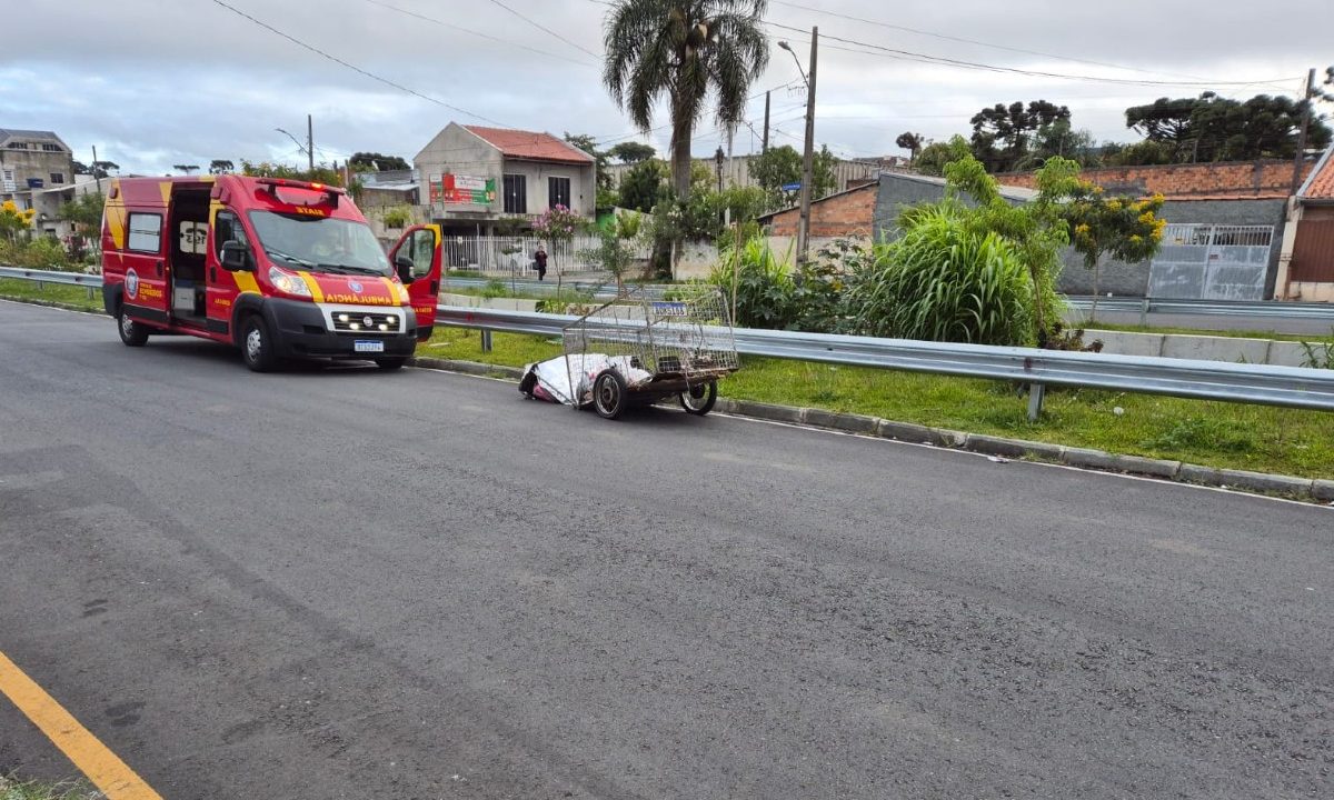 Carrinho de recicláveis ao lado de ambulância do Siate marca o local onde uma catadora de recicláveis foi encontrada morta em rua de Curitiba; o corpo está coberto por manta térmica na via asfaltada, com casas ao fundo.
