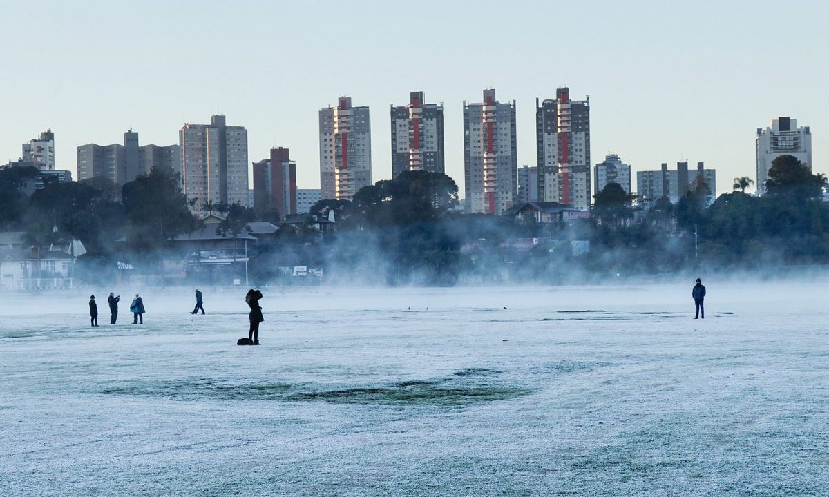 Imagem mostra o Parque Barigui em Curitiba com geada. O gramado está coberto. Atrás aparecem os prédios. Com o clima mudando, geadas não são tão comuns mais.