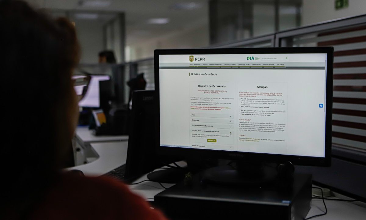 Imagem mostra mulher de costas, olhando para tela do computador. A tela mostra como registrar um boletim de ocorrência pela internet, totalmente online