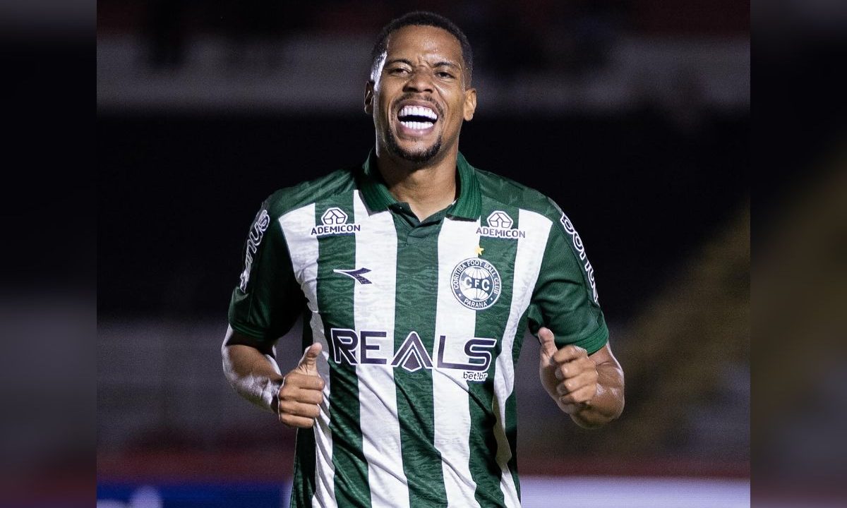 Iury Castilho aparece em primeiro plano, vestindo a camisa verde e branca do Coritiba. O atacante está sorrindo, com a boca aberta, e faz um gesto com as duas mãos levantadas próximas ao peito. O fundo da imagem está desfocado, indicando um ambiente de jogo noturno.