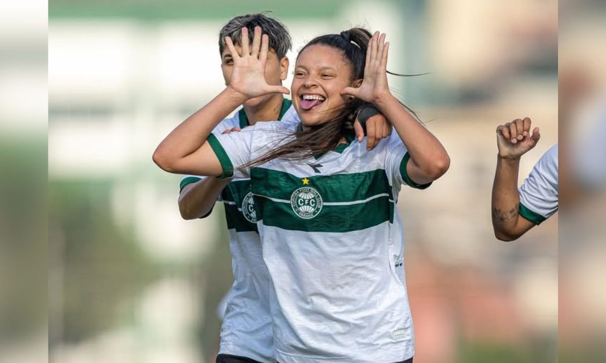 Jogadora do Coritiba feminino aparece em primeiro plano comemorando um gol. Ela veste uniforme branco com uma faixa horizontal verde no peito e o escudo do clube centralizado. Com um sorriso aberto e expressão de alegria, levanta as duas mãos próximas ao rosto, com os dedos abertos, em gesto de comemoração. Ao fundo, outra companheira de equipe também comemora, parcialmente encoberta, com o braço sobre o ombro da atleta em destaque. A imagem tem fundo desfocado, sugerindo um campo de futebol, e transmite um momento de celebração durante a partida.