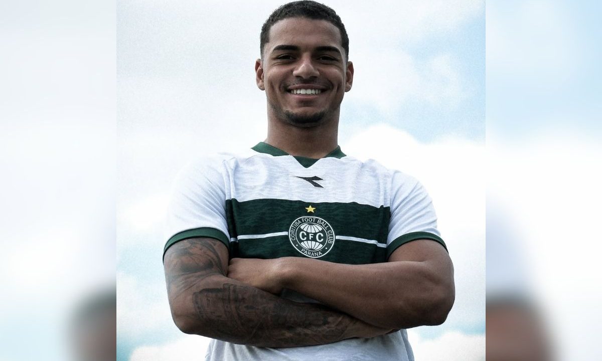 Na foto está Renato Marques. Homem jovem, de aproximadamente 22 anos, com pele morena clara e cabelo curto escuro, aparece em primeiro plano com expressão sorridente. Ele está de braços cruzados, olhando diretamente para a câmera, transmitindo confiança. Veste a camisa branca do Coritiba com uma faixa horizontal verde no peito e o escudo do clube centralizado. Seus braços têm tatuagens visíveis. O fundo é um céu claro com algumas nuvens, desfocado, destacando o jogador como elemento principal da imagem.