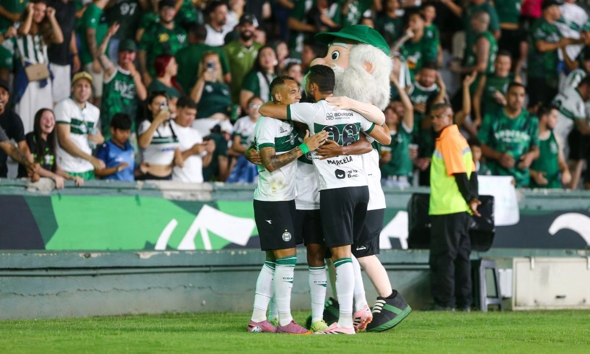 Jogadores do Coritiba se abraçam em comemoração de gol no gramado do Couto Pereira, com Pedro Rocha no centro do grupo, próximos da torcida alviverde ao fundo, que aparece em pé e vibrando nas arquibancadas.