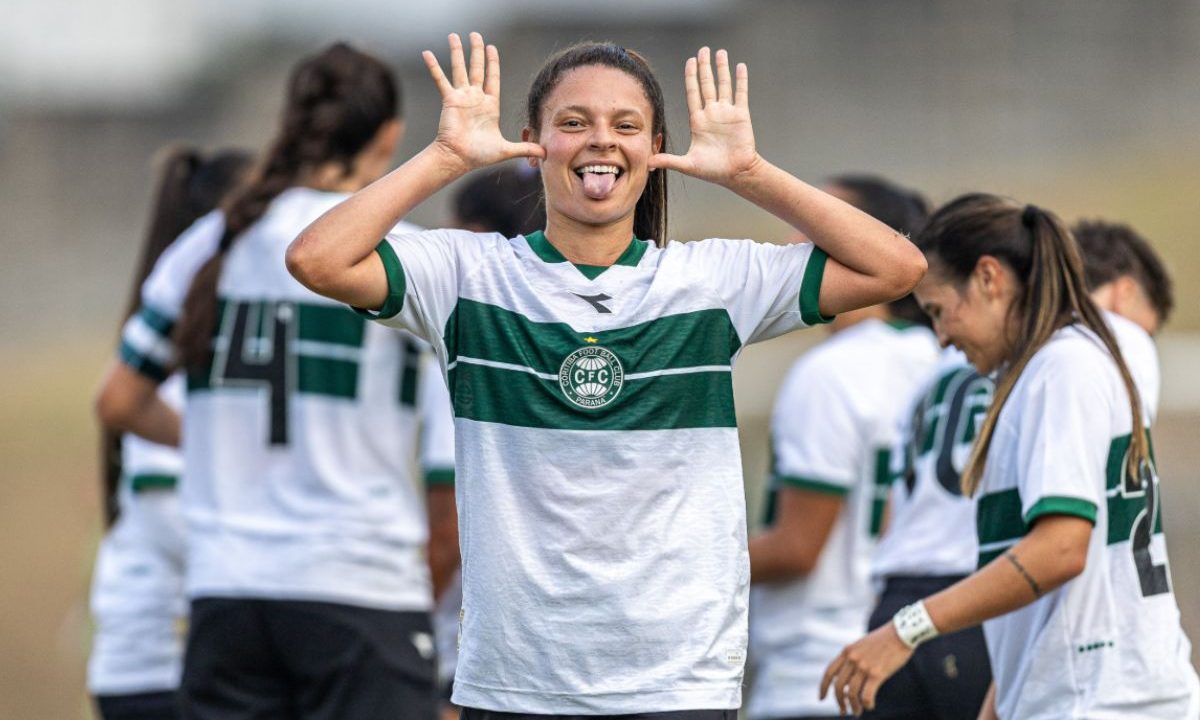 Em primeiro plano, uma jogadora do Coritiba aparece sorrindo e comemorando, olhando diretamente para a câmera. Ela veste o uniforme branco com faixa horizontal verde no peito e o escudo do clube centralizado. Com expressão descontraída, levanta as duas mãos ao lado da cabeça, com os dedos abertos, e coloca a língua para fora em tom de brincadeira. Ao fundo, outras atletas do time, também uniformizadas, aparecem desfocadas, algumas de costas e outras em movimento, sugerindo um momento de celebração ou descontração durante a partida ou treino.