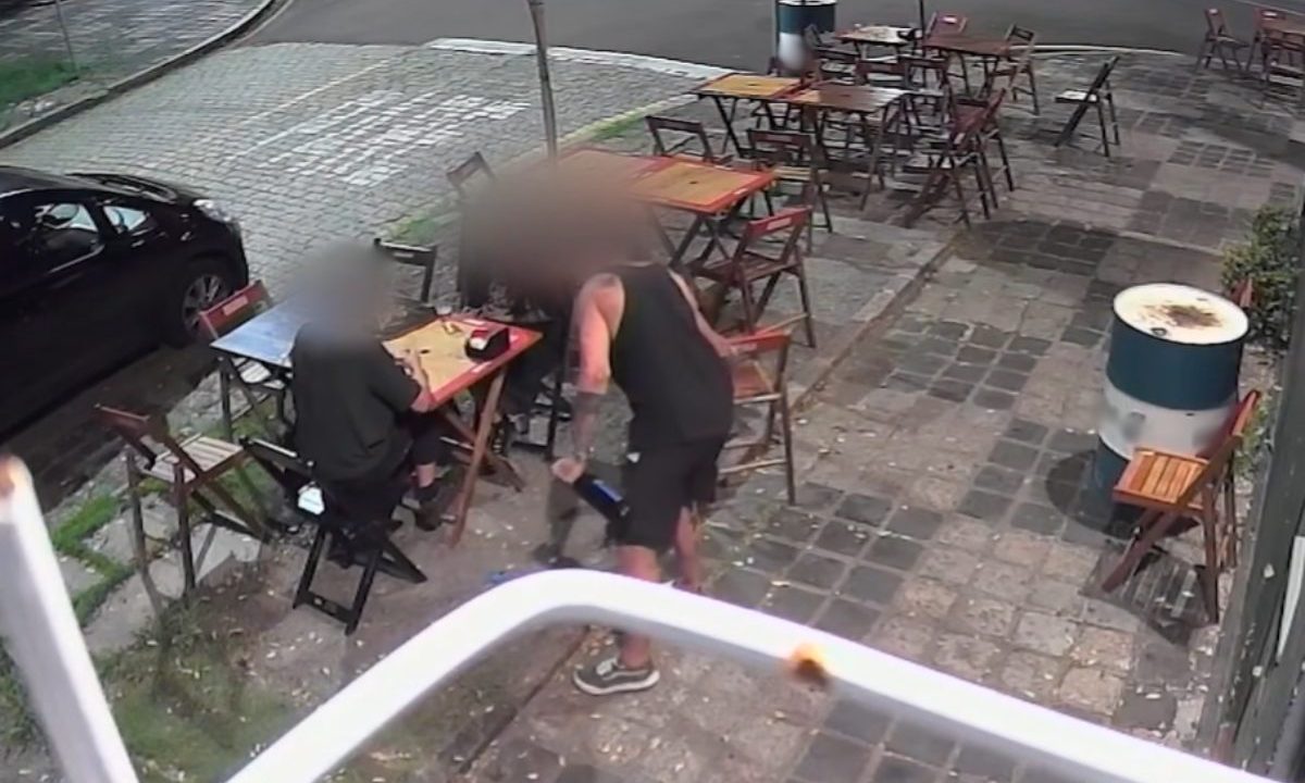 Imagem mostra um casal sentado em uma mesa de bar. O suposto cozinheiro acusado da agressão está em pé ao lado do homem. Ele usa uma regata preta e tem uma garrafa de vidro na mão. O casal parece tranquilo. Eles usam roupas pretas. Na calçada, há diversas mesas e cadeiras.