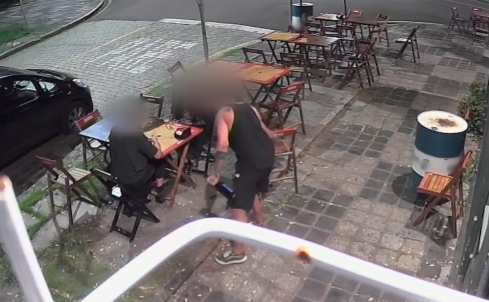 Imagem mostra um casal sentado em uma mesa de bar. O suposto cozinheiro acusado da agressão está em pé ao lado do homem. Ele usa uma regata preta e tem uma garrafa de vidro na mão. O casal parece tranquilo. Eles usam roupas pretas. Na calçada, há diversas mesas e cadeiras.
